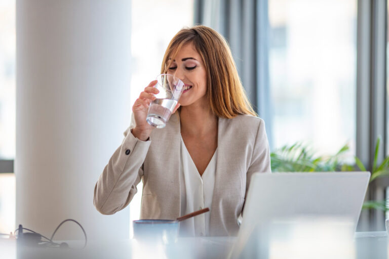 Trinkwasserversorgung am Arbeitsplatz - eine Frau am Tisch trinke aus einem Gals mit Wasser